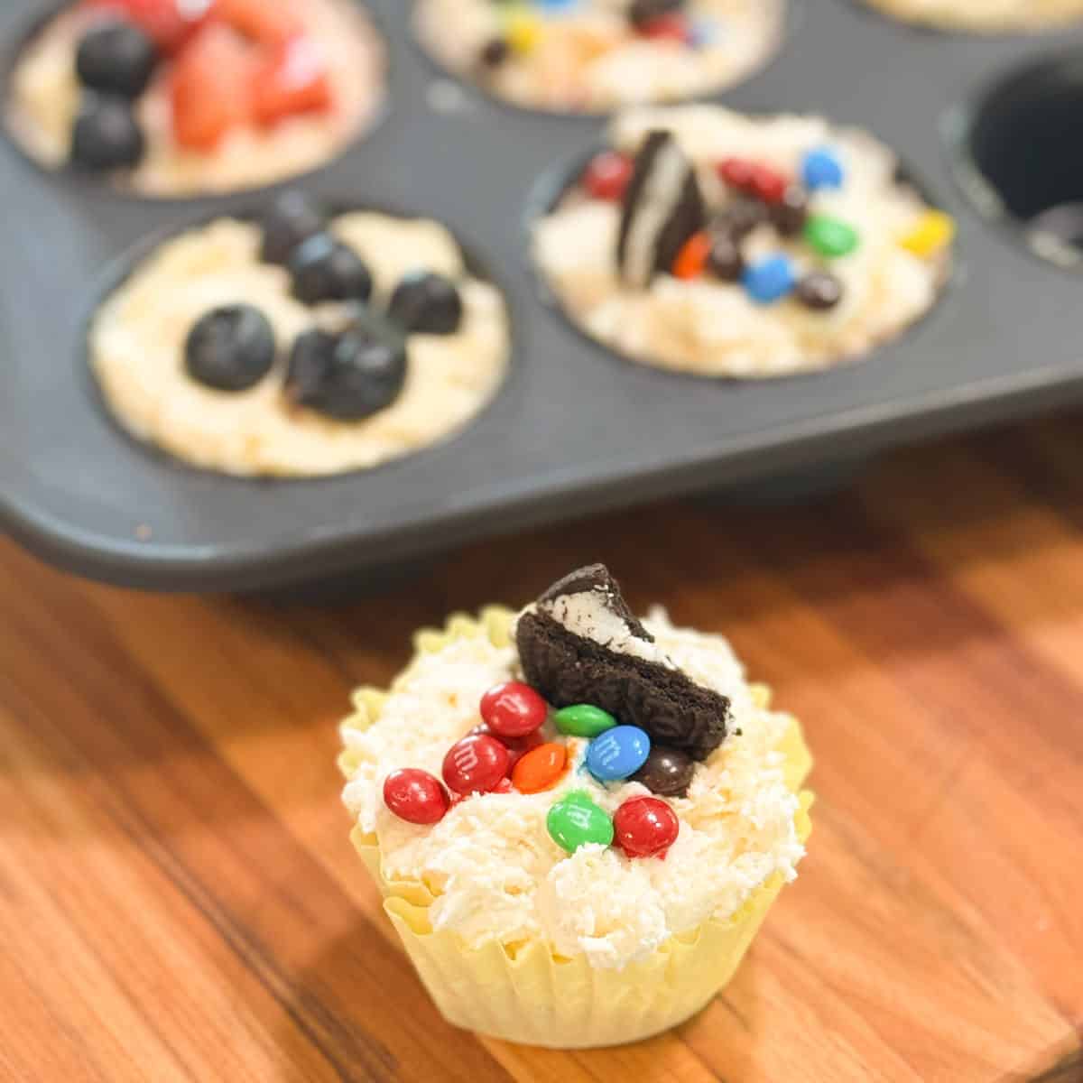 A muffin tin holds unbaked cupcake batter topped with strawberries, blueberries, Oreo pieces, and M&M candies. In the foreground, a single cupcake in a yellow wrapper is decorated with M&Ms and an Oreo piece.