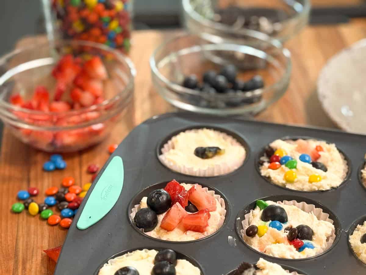 A muffin tin with batter in paper liners is topped with blueberries, strawberries, and colorful candy pieces. Bowls of strawberries, blueberries, chocolate sandwich cookies, and mini candy-coated chocolates are in the background.