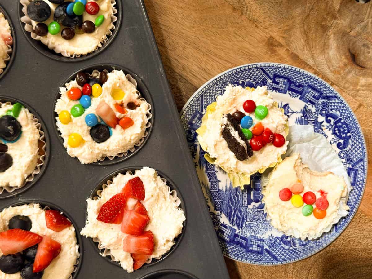 A muffin tin with six cheesecake cupcakes topped with fruit and candy sits beside a blue and white plate holding two more decorated cheesecake cupcakes on a wooden surface.