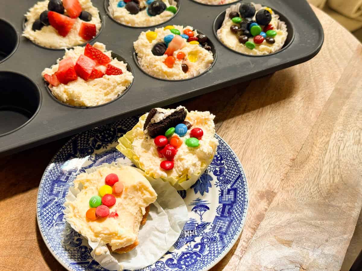 A muffin tin with six cheesecake cupcakes topped with assorted candies, fruits, and cookies sits on a wooden surface. Two finished cupcakes, unwrapped and topped with M&M's and Oreo pieces, are on a blue and white plate.