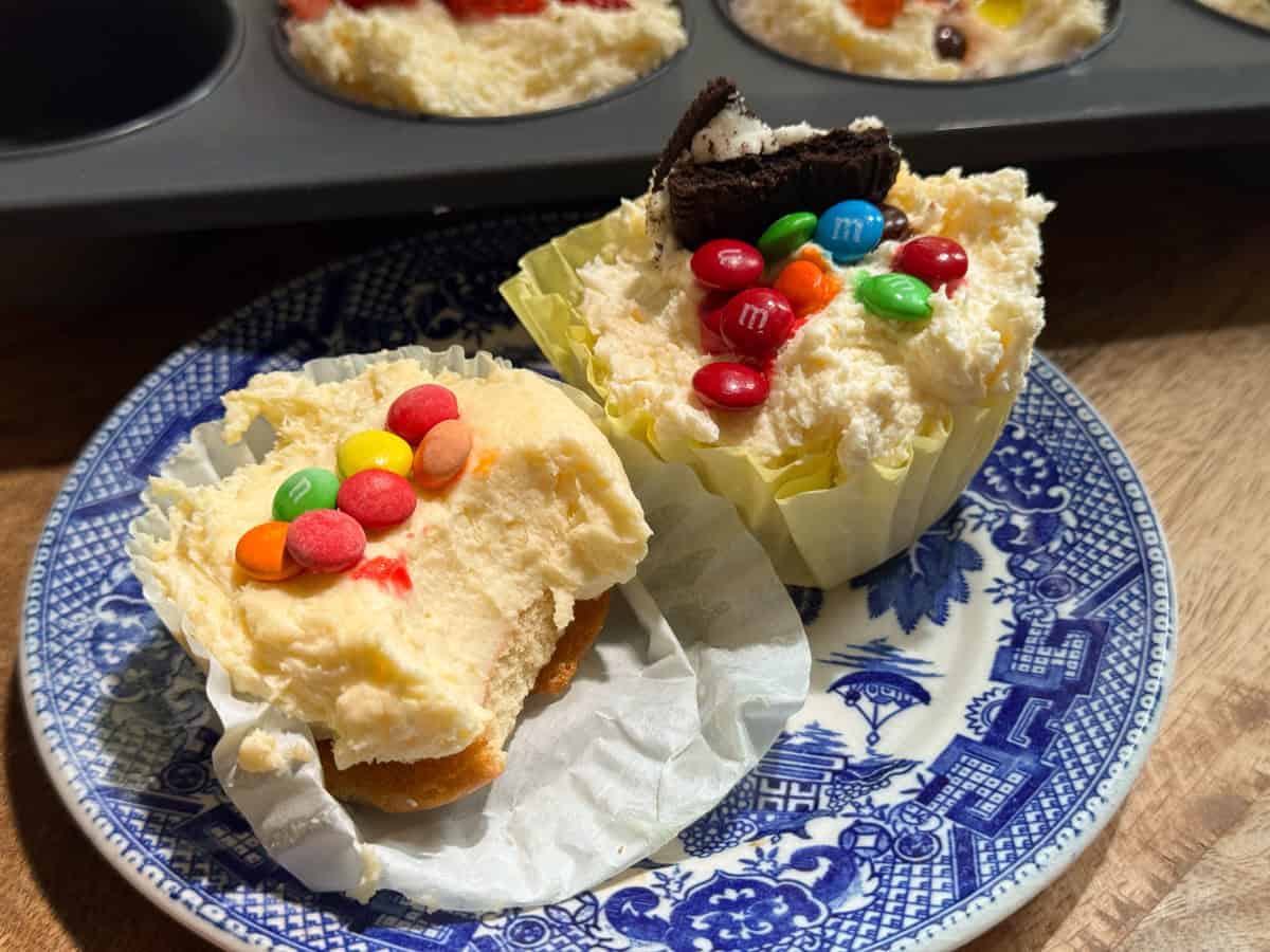 Two mini cheesecakes on a blue and white patterned plate, each topped with colorful M&M candies; one also has a piece of chocolate cookie. The cheesecakes are in yellow paper liners, with a baking tray in the background.
