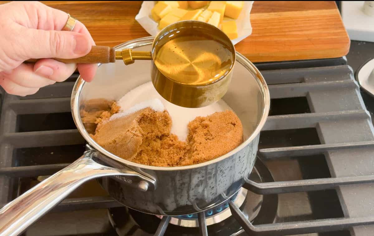 A hand holds a measuring cup of liquid above a saucepan containing brown sugar and white sugar on a stove, with a plate of butter in the background.