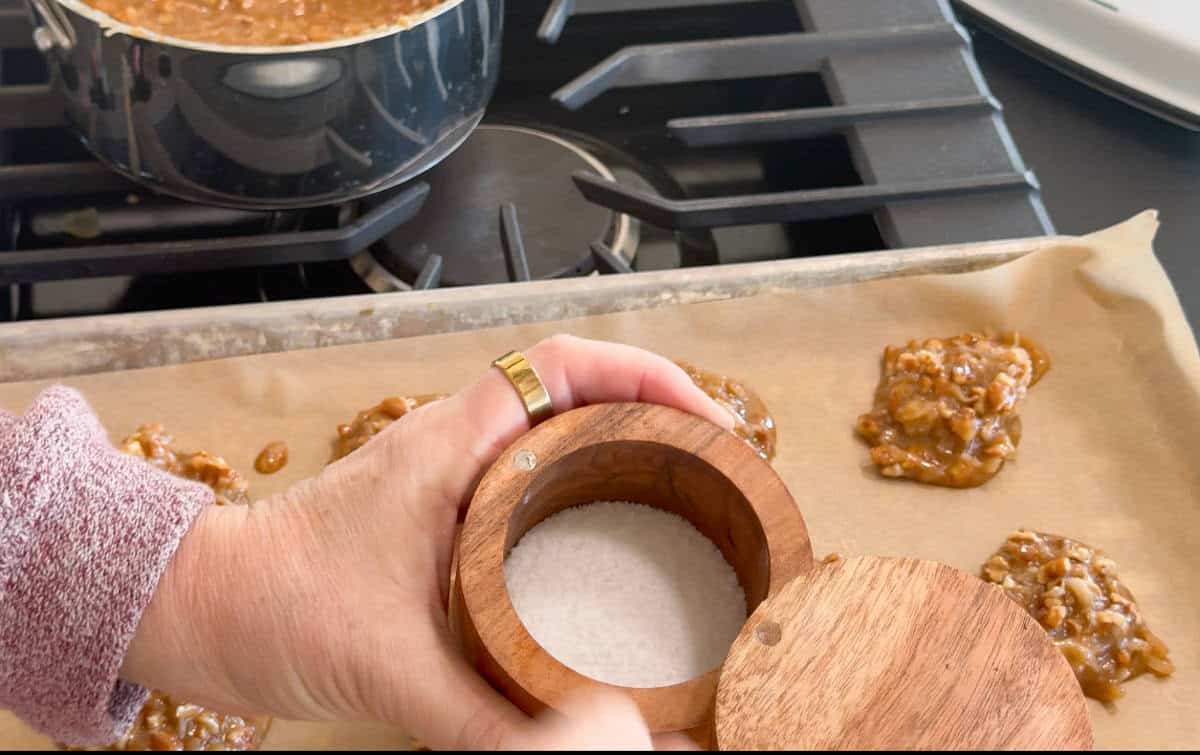 A person holds an open wooden salt cellar above a baking sheet with clusters of cookie dough on parchment paper, in front of a stovetop with a pot of mixture simmering.