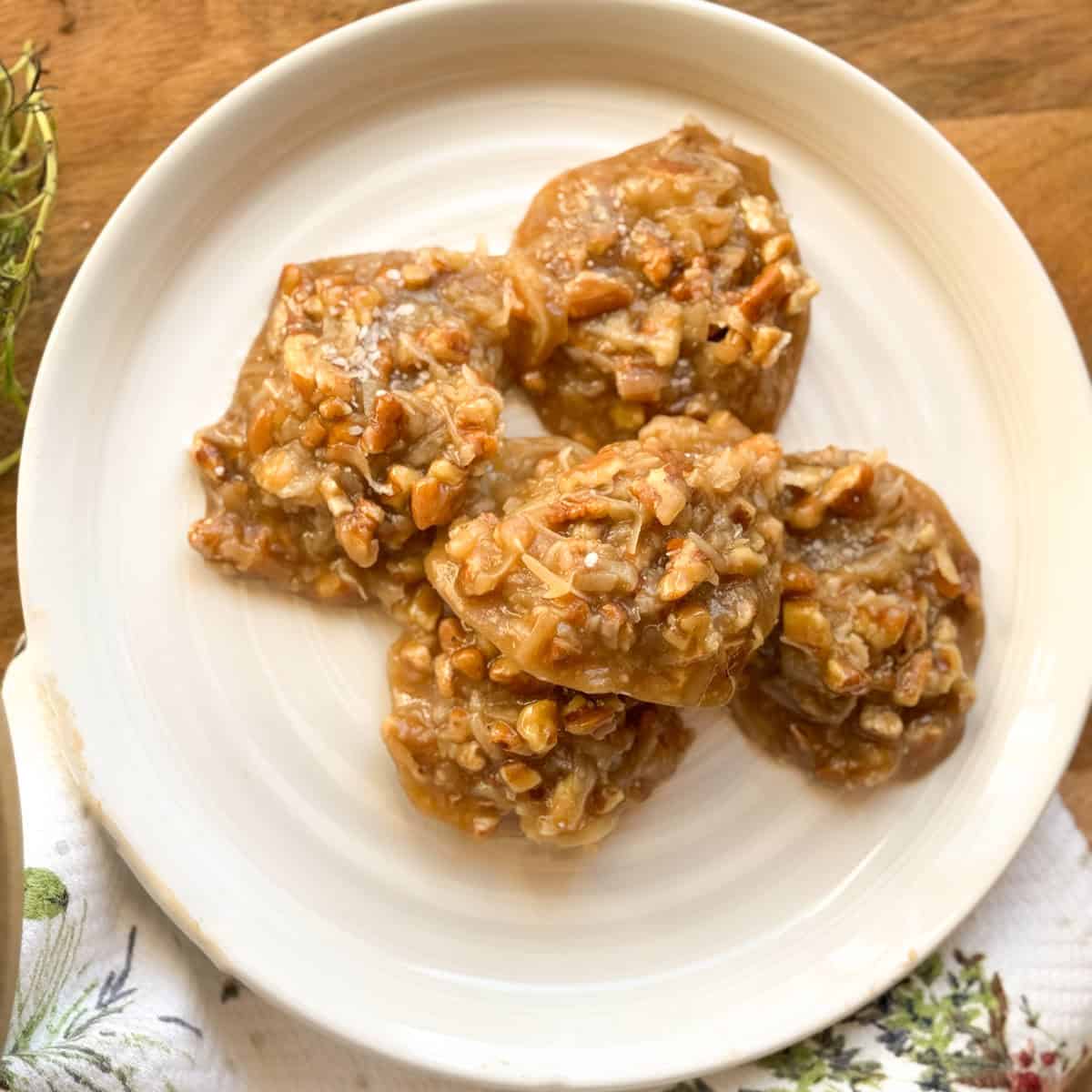 Five pecan praline cookies are arranged on a white plate. The cookies appear caramelized and contain visible pecan pieces. The plate is placed on a wooden surface next to a patterned cloth.