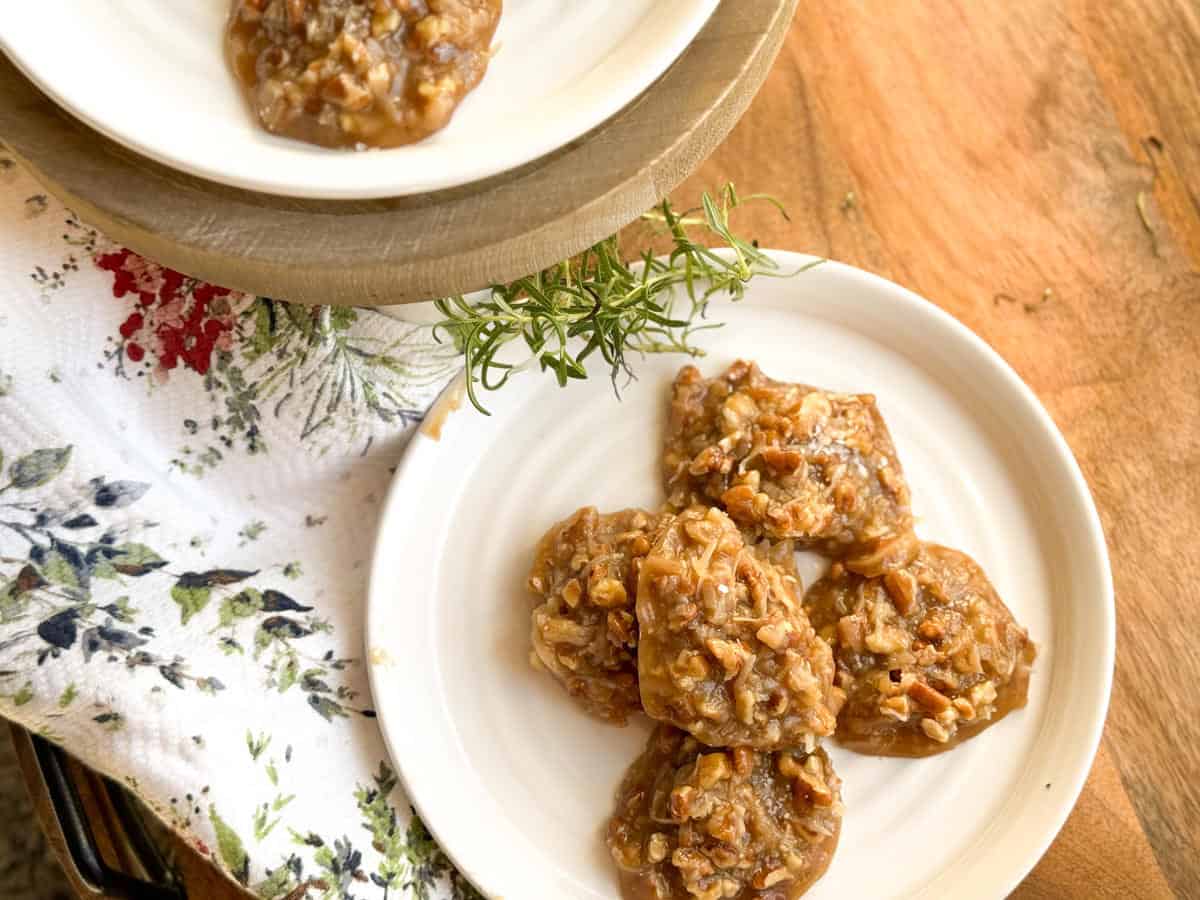 Five praline cookies with a caramel and nut appearance are arranged on a white plate placed on a wooden surface, next to a floral-patterned cloth and a sprig of fresh rosemary. Another plate is partially visible above.