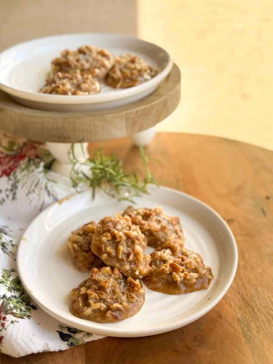 Two white plates with pecan praline cookies are placed on a wooden surface. One plate is elevated on a wooden stand, and a sprig of greenery rests beside them. A patterned cloth is partially visible on the left.