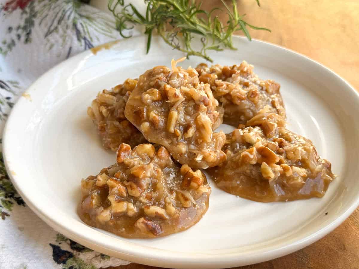 A white plate holds several pecan pralines with a glossy, caramel-like coating and pieces of chopped pecans. A sprig of rosemary and a patterned cloth are in the background.
