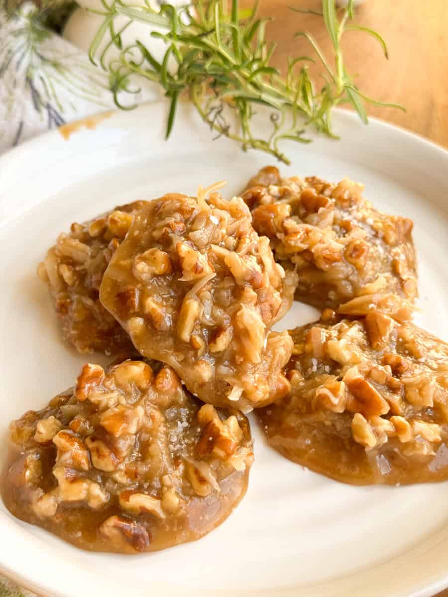 Five praline cookies topped with chopped pecans are arranged on a white plate. There is a sprig of fresh rosemary and part of a patterned napkin visible in the background.