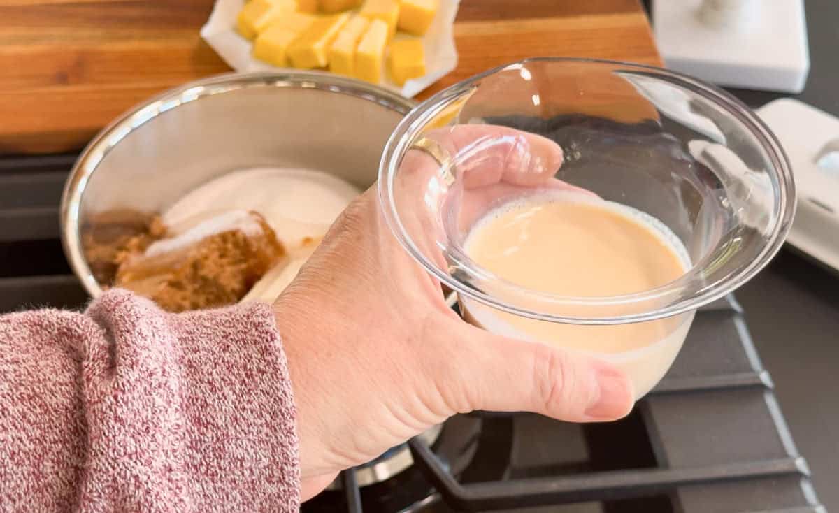 A hand holds a small clear bowl of cream above a saucepan containing brown sugar and white sugar on a stove, with cubed butter on a cutting board in the background.
