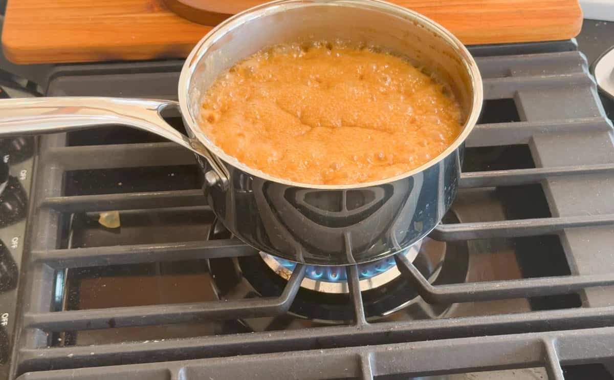 A stainless steel saucepan with a caramel-colored mixture boiling on a gas stove burner, with bubbles covering the surface of the liquid. The stove grate and part of a wooden cutting board are visible.