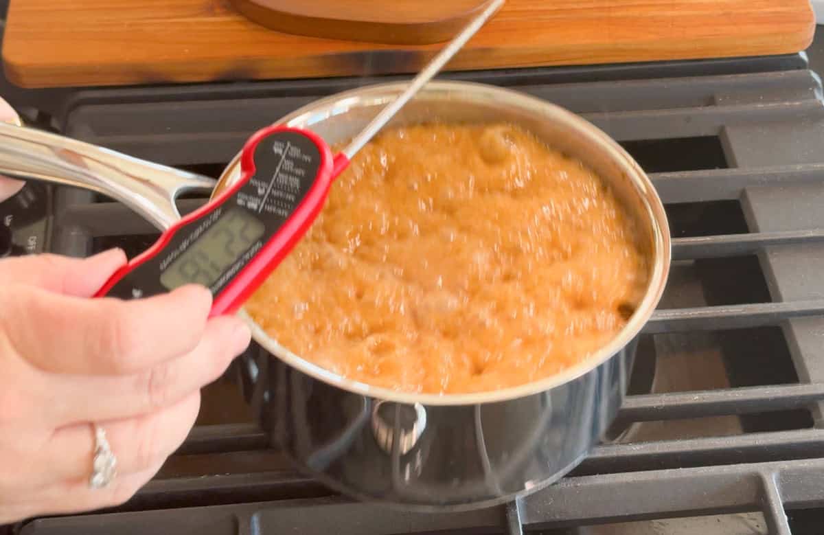 A person holds a digital thermometer over a saucepan of bubbling caramel on a stovetop, measuring the temperature of the mixture.