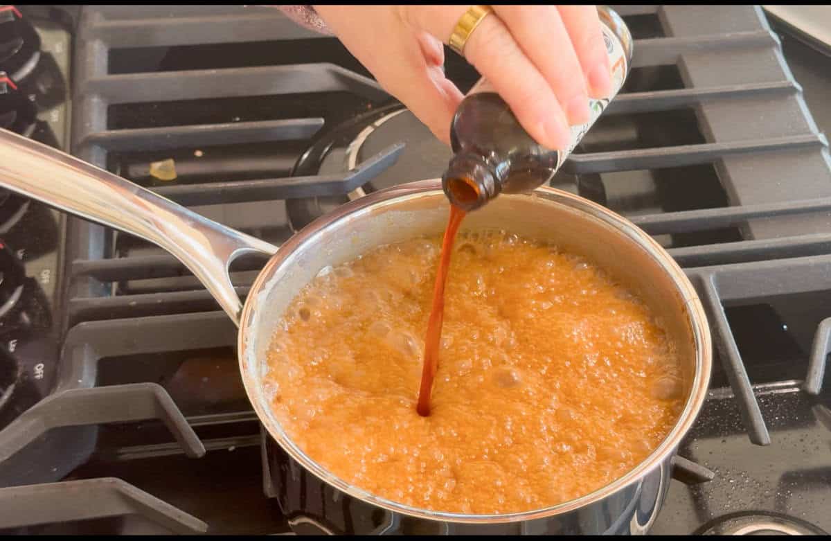 A hand pours liquid vanilla extract from a bottle into a saucepan with bubbling caramel on a stove.
