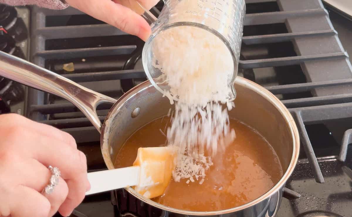 A hand pours shredded coconut from a measuring cup into a saucepan of brown liquid on a stove, while another hand stirs with a spatula.