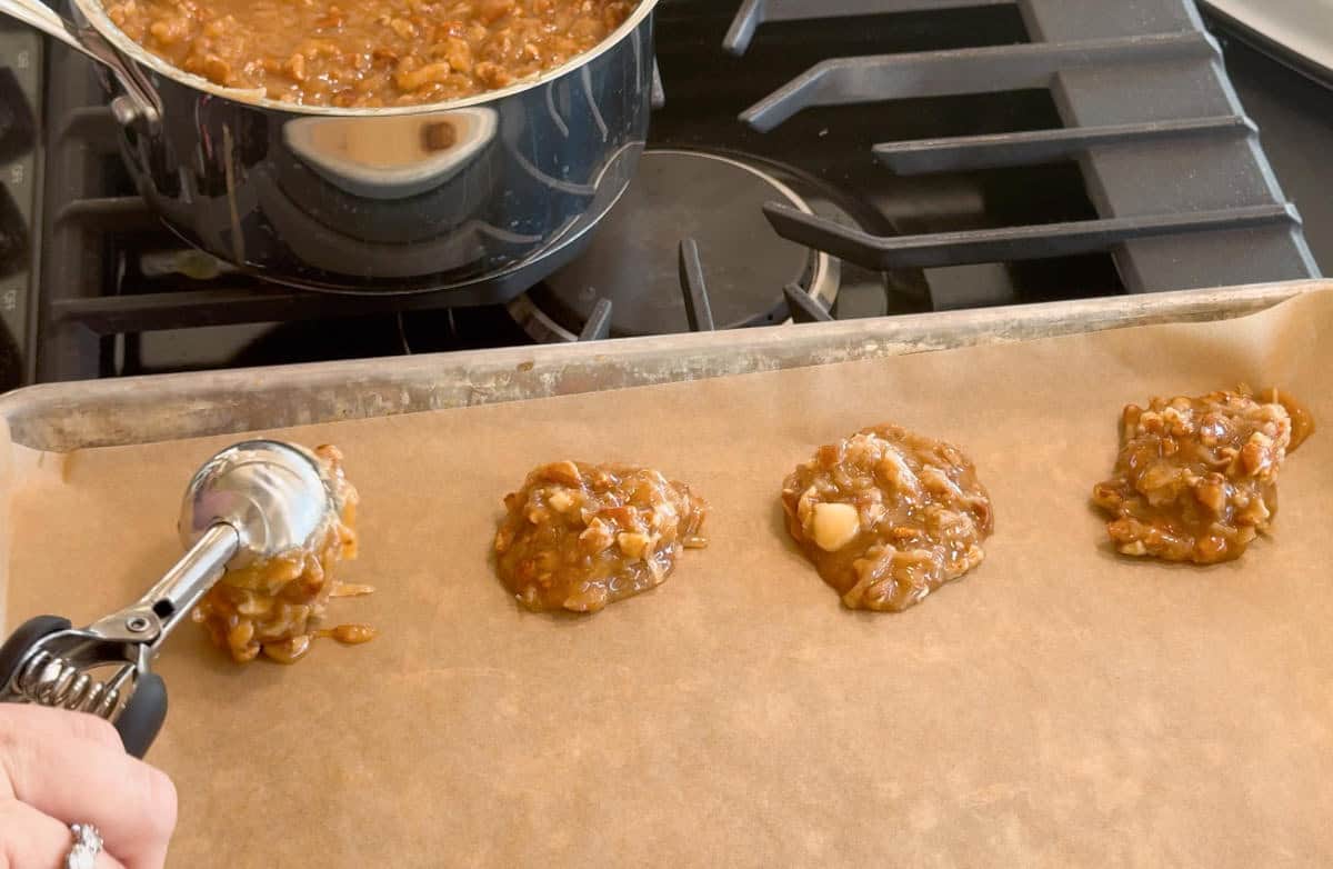 A hand uses a metal scoop to place portions of raw cookie dough onto a parchment-lined baking sheet; a pot of dough sits on the stove in the background.