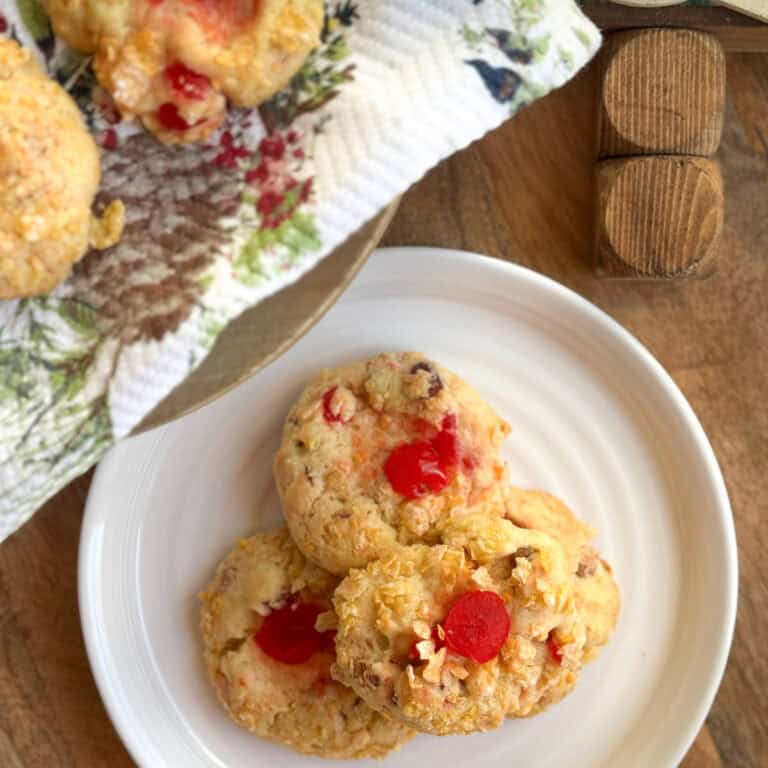 Three cookies with red cherry pieces and visible nuts are arranged on a white plate. More cookies are on a festive napkin with a pinecone design, displayed on a wooden surface.