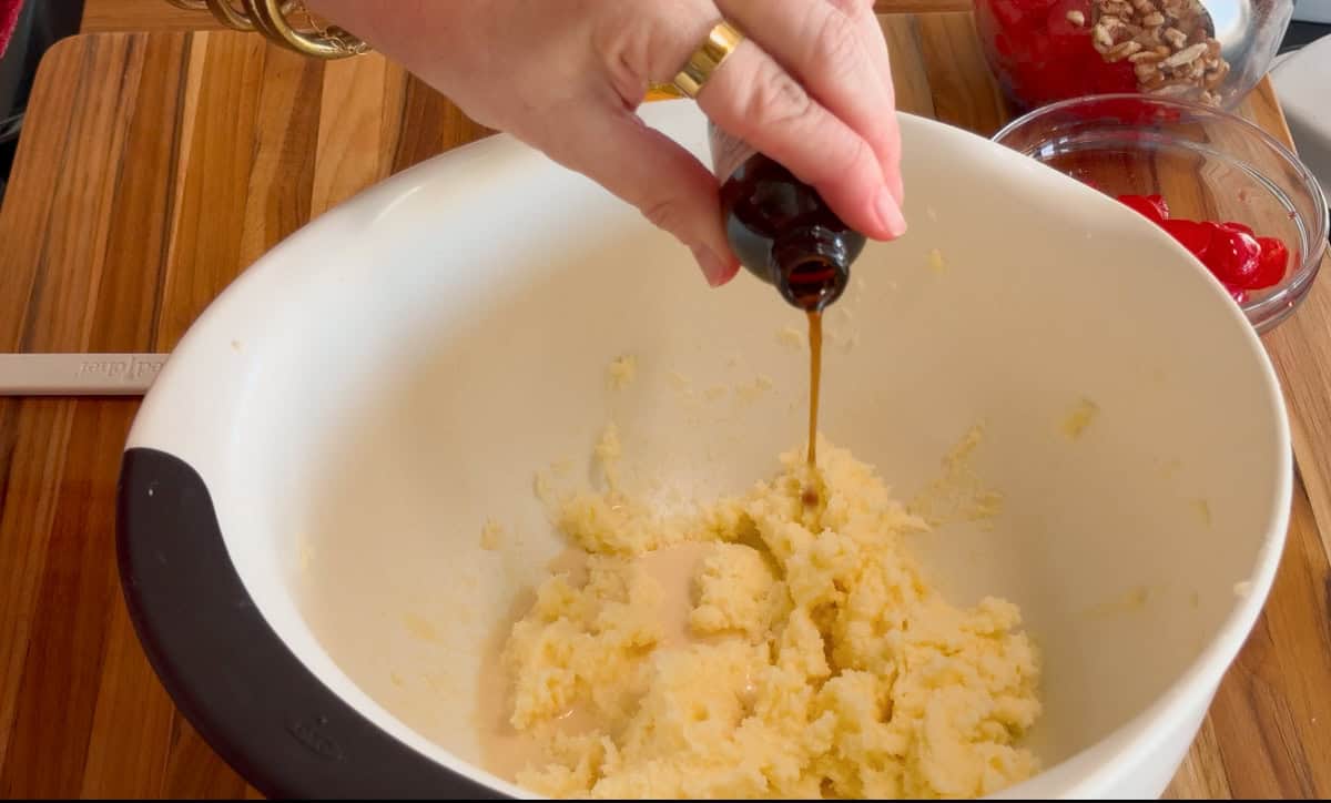 A hand is pouring vanilla extract from a bottle into a bowl of creamed sugar and butter. The bowl is on a wooden surface, with small bowls of pecans and cherries in the background.
