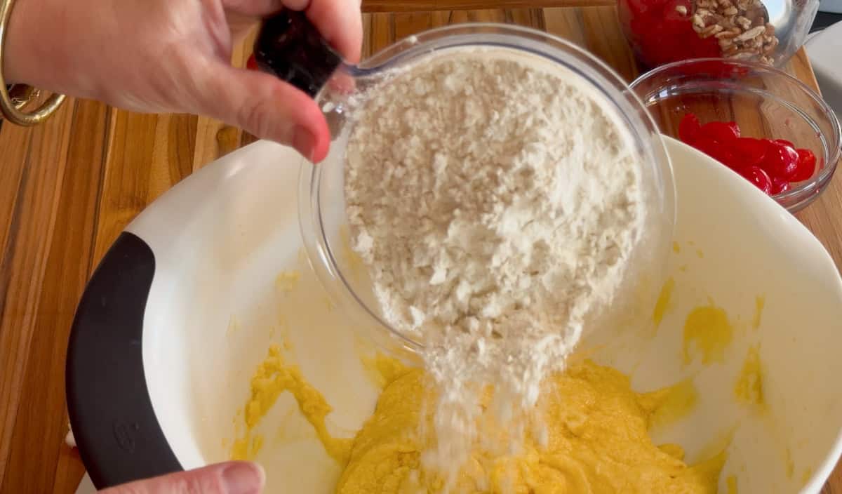A person pours a cup of flour into a mixing bowl containing a yellow batter. Other ingredients, including cherries and nuts, are visible in small bowls nearby on the wooden counter.