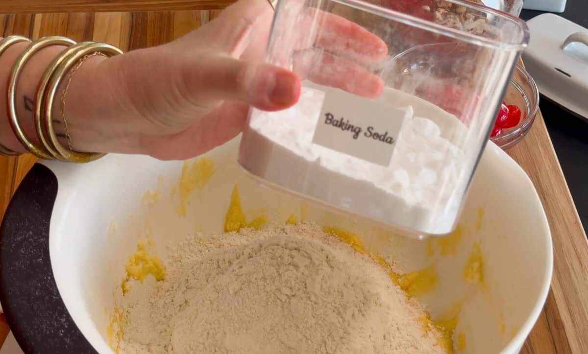 A hand holding a clear container labeled "Baking Soda" above a mixing bowl with flour and a yellow mixture, on a wooden countertop. Gold bangles are on the wrist.