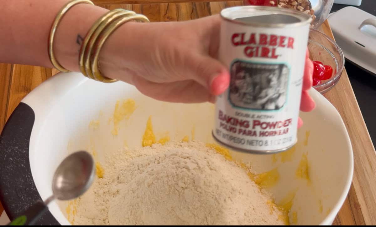 A hand holding a can of Clabber Girl baking powder above a white mixing bowl filled with flour; a spoon and other baking ingredients are visible in the background on a wooden surface.