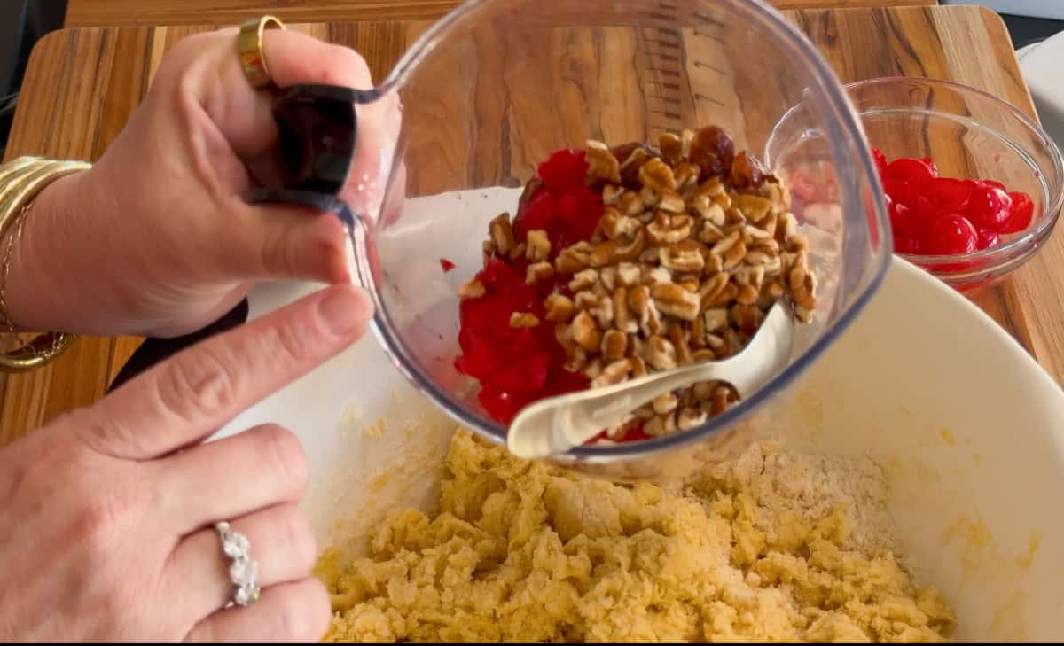 A person holds a clear measuring cup filled with chopped pecans and red candied cherries over a mixing bowl containing cookie dough. A spoon rests in the cup. A bowl of maraschino cherries sits in the background.
