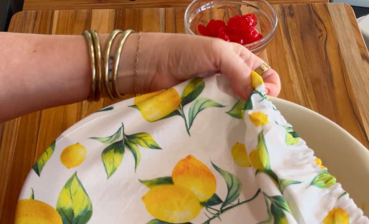 A hand wearing several bangles holds a white cloth with a lemon pattern over a bowl. In the background, there is a small glass bowl containing red cherries on a wooden surface.