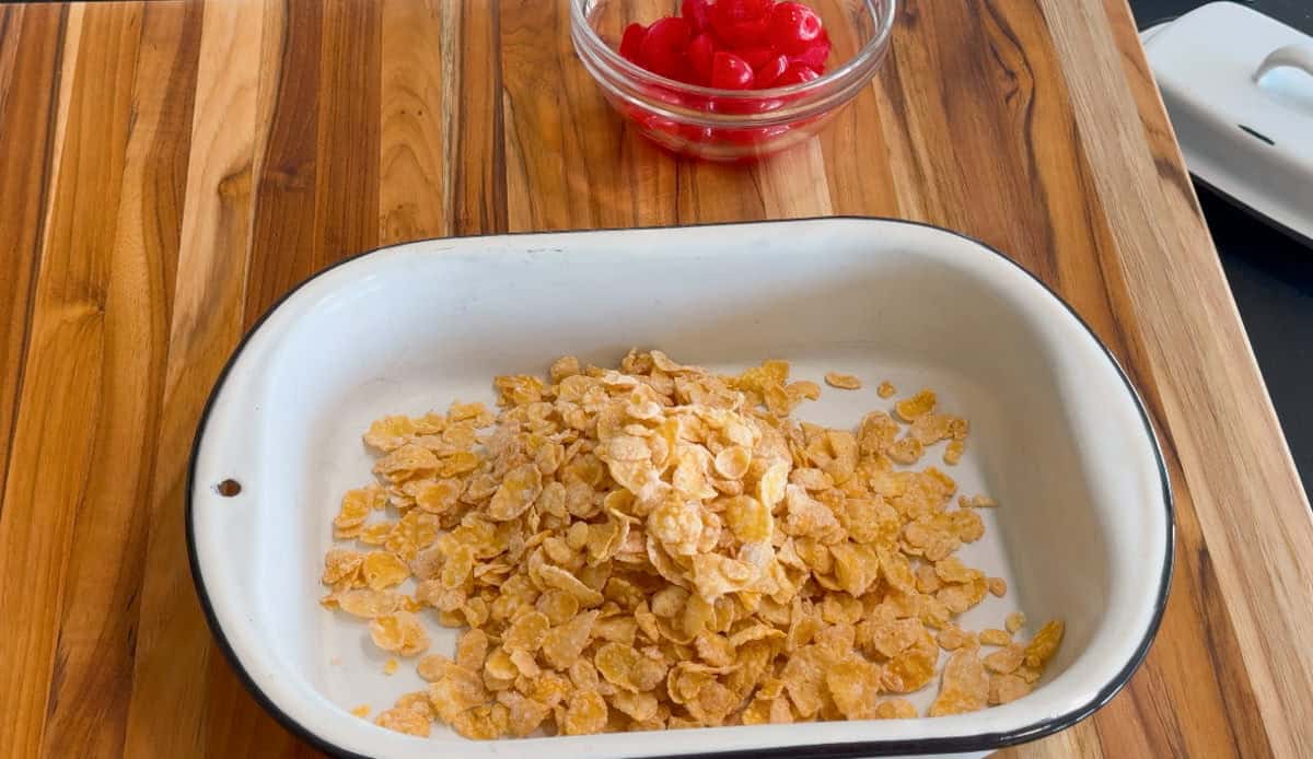A white baking dish filled with cornflakes sits on a wooden surface. Behind it, there is a small glass bowl containing red maraschino cherries.