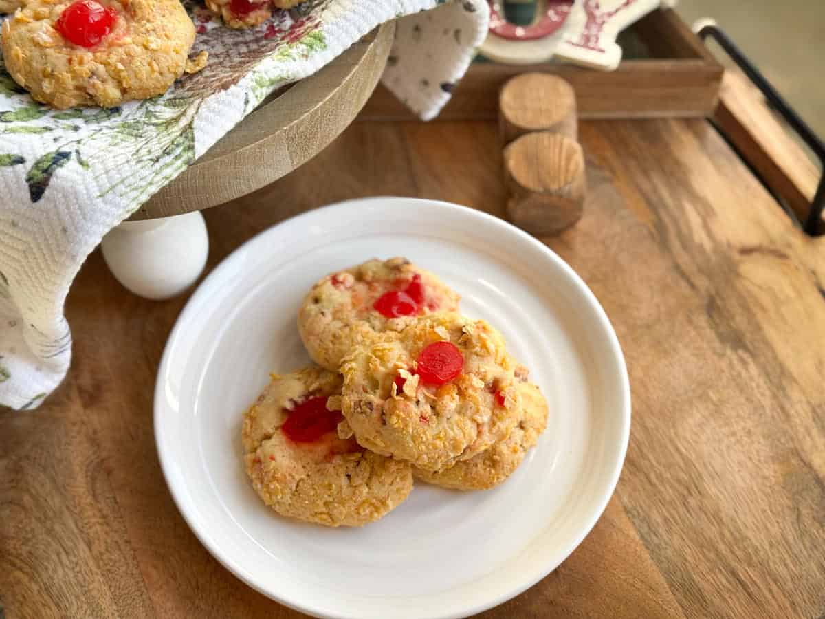 Three cookies with pieces of red cherry on top are arranged on a white plate. More cookies are partially visible on a raised tray covered with a cloth. The cookies and tray are on a wooden surface.