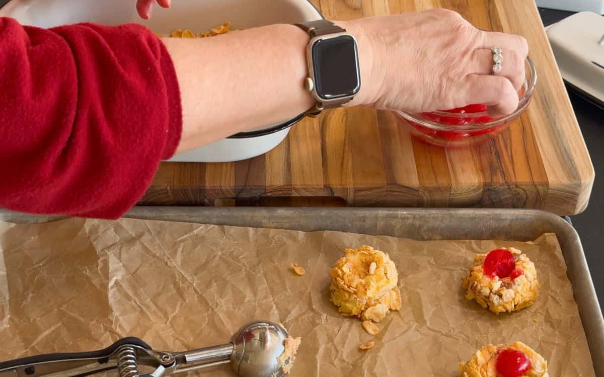 A person wearing a red sleeve and a smartwatch places a red cherry on top of a cookie dough ball on a parchment-lined baking sheet. Other cookie dough balls and a metal scoop are also visible.