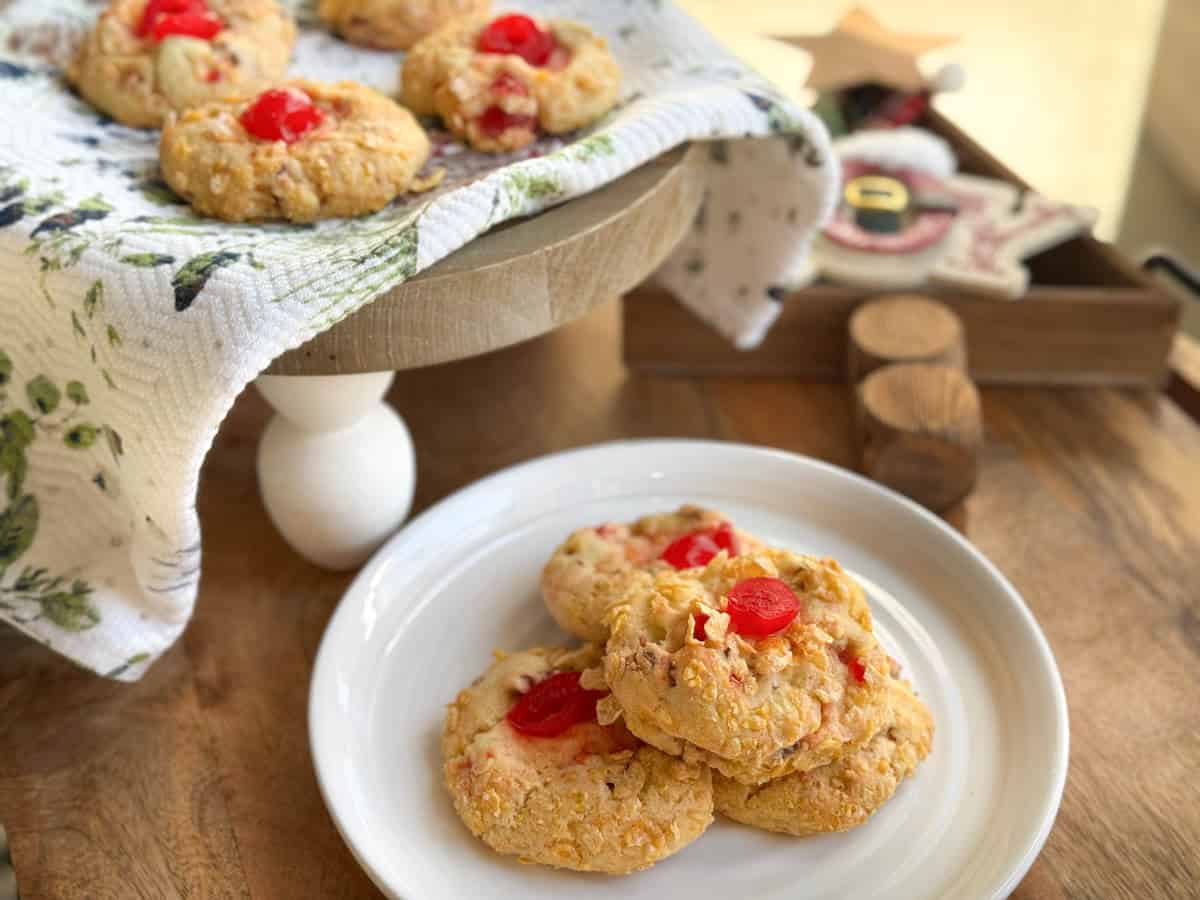 A white plate with three cookies topped with red cherries sits on a wooden table. Behind it, more cherry-topped cookies rest on a cloth-covered cake stand. A decorative tray with ornaments is in the background.