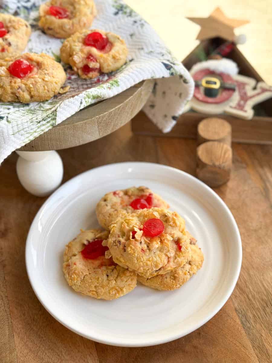 Three cookies with red cherry centers are on a white plate in the foreground. In the background, more cookies rest on a raised wooden tray covered with a cloth. Holiday-themed decorations are partially visible.