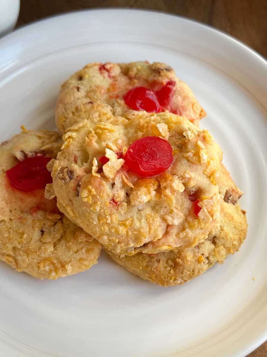 Three cookies with a golden, crumbly texture and pieces of red cherry on top are stacked on a white plate. The cookies have small bits and chunks visible throughout.