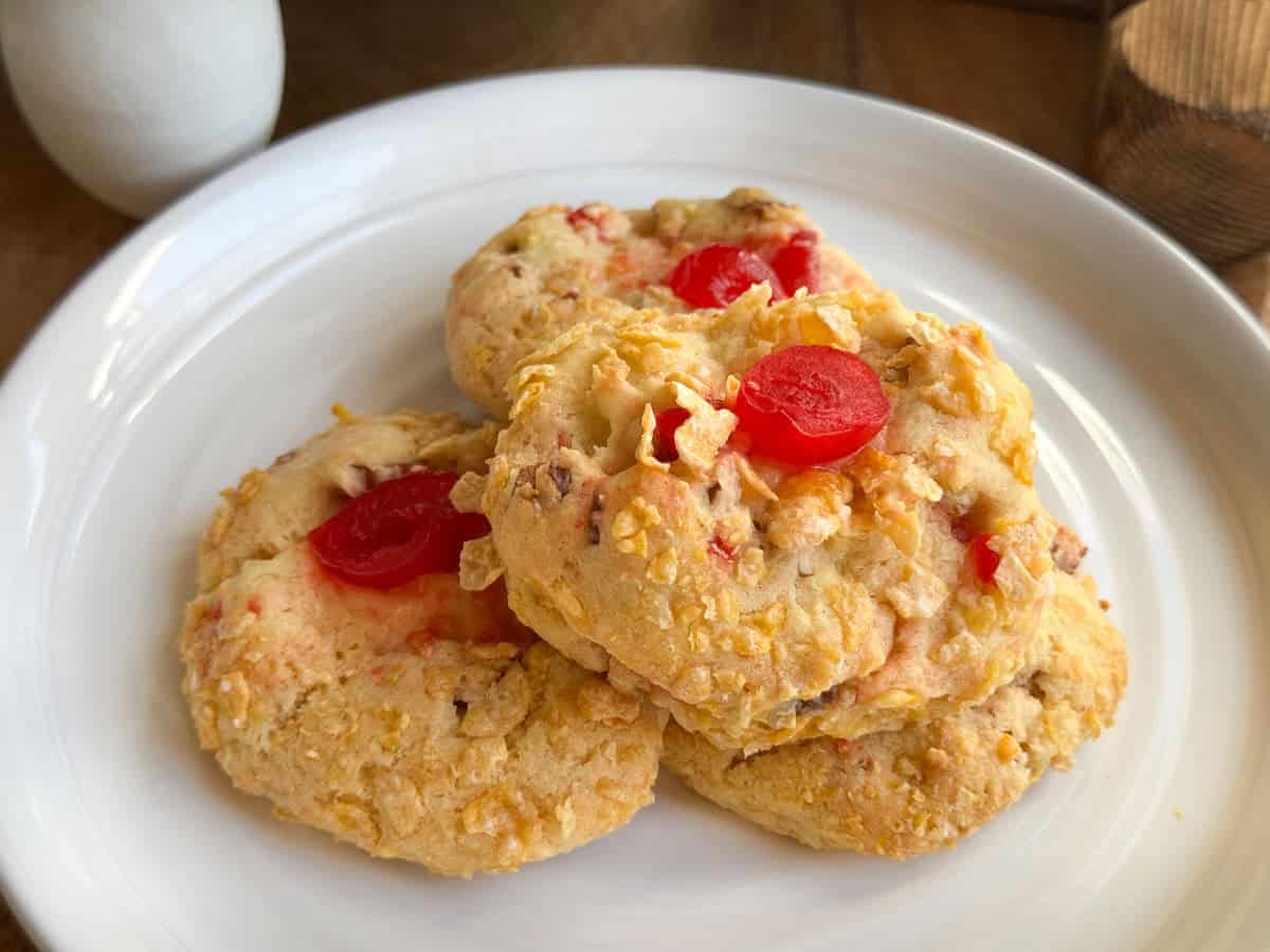 Four golden cookies topped with red candied cherries are stacked on a white plate. The cookies have a crumbly texture and visible bits of cherries and nuts. The plate sits on a wooden surface.