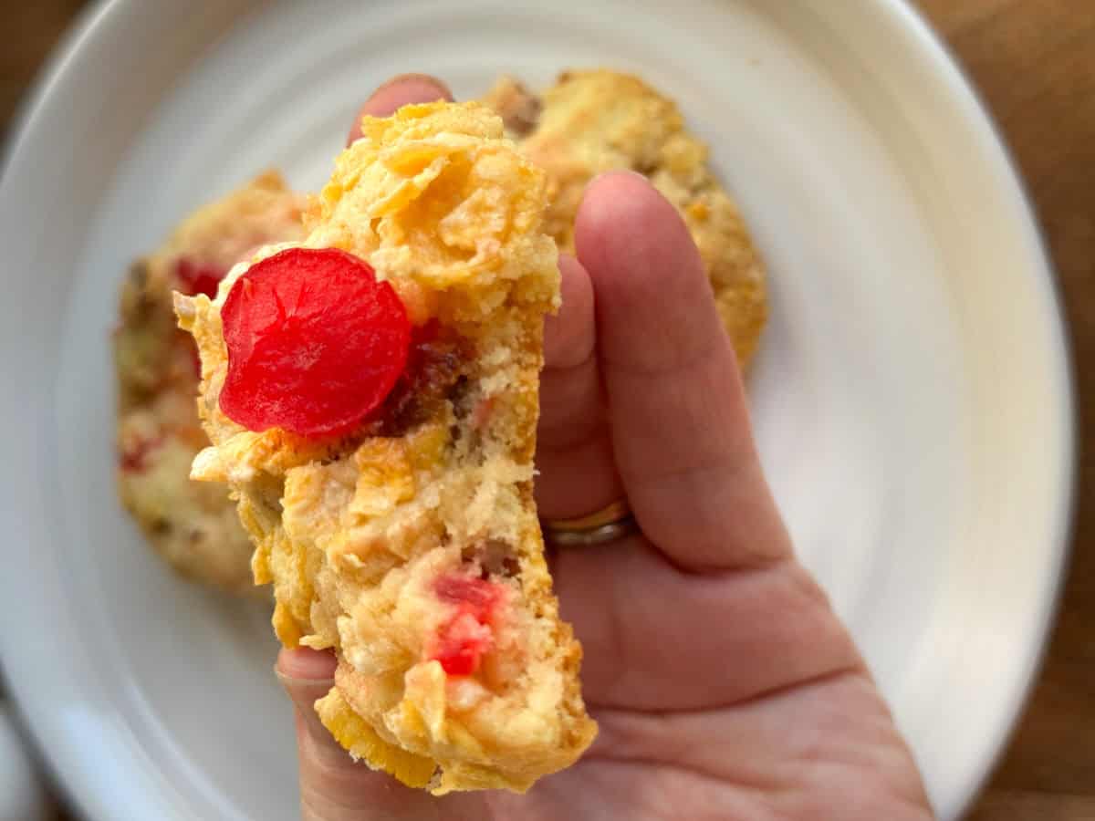 A hand holding up a cookie with a bright red cherry and visible chunks, over a white plate with another cookie in the background.