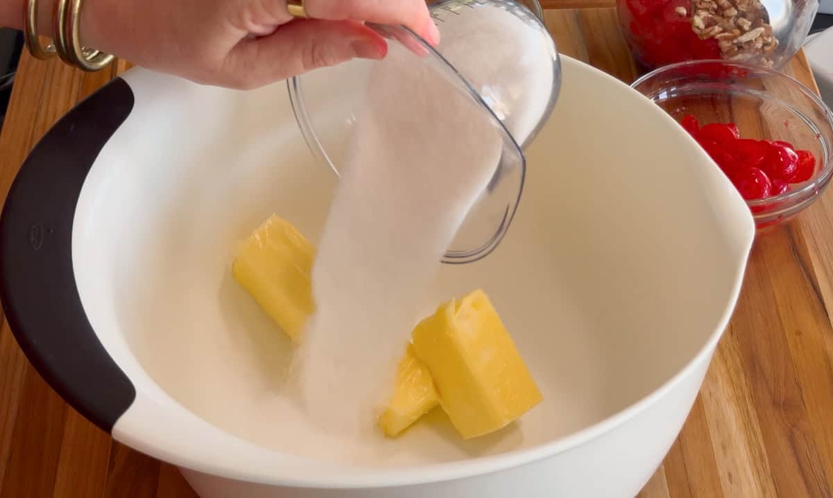 A hand pours granulated sugar from a measuring cup into a white mixing bowl containing two sticks of butter. A glass bowl with red cherries and another with nuts are in the background on a wooden surface.