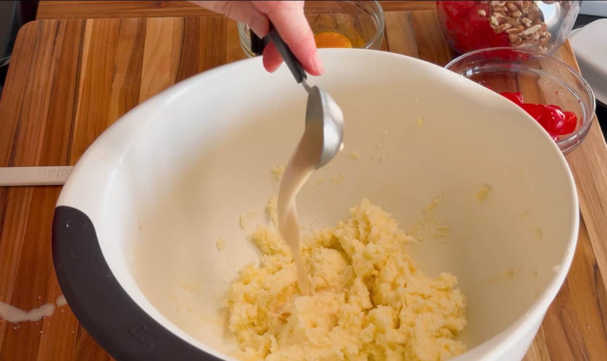 A hand pours liquid from a metal spoon into a white mixing bowl containing a creamed mixture. Surrounding the bowl on a wooden surface are an egg, chopped nuts, and red cherries in small glass bowls.