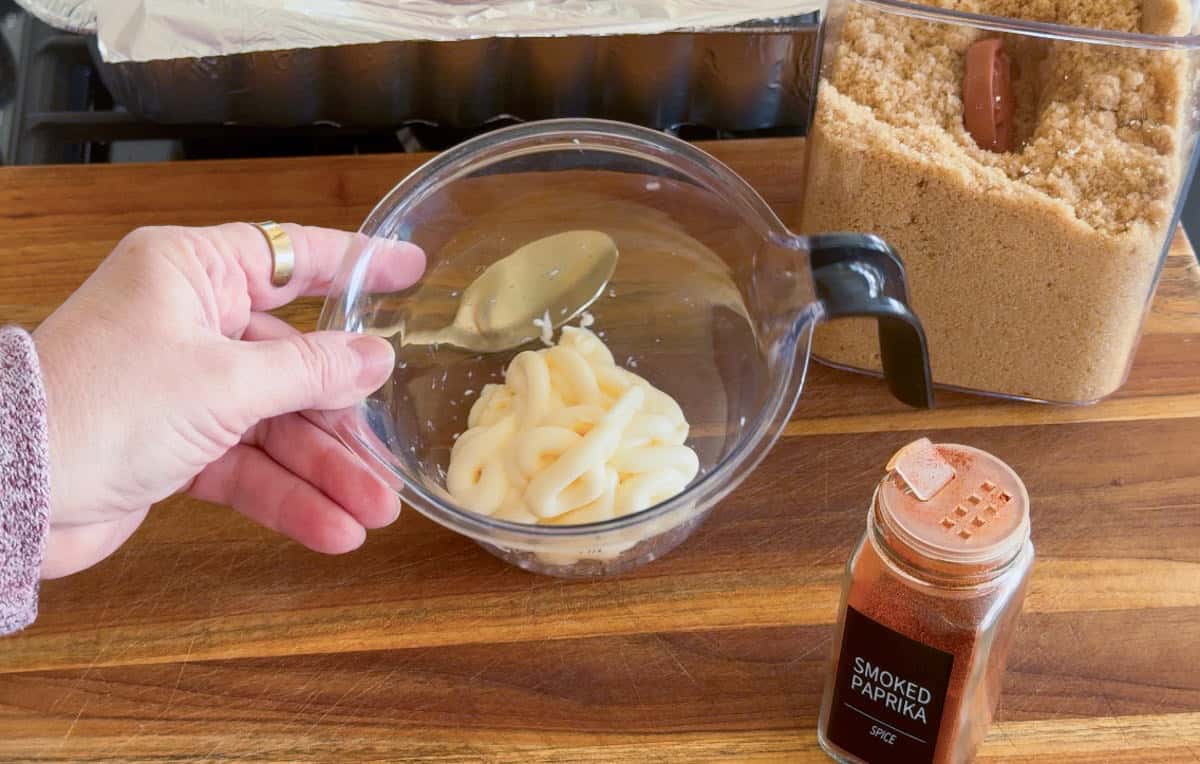 A hand holds a measuring cup containing mayonnaise above a wooden surface. Nearby are a container of brown sugar, a jar of smoked paprika, and a foil-lined pan in the background.