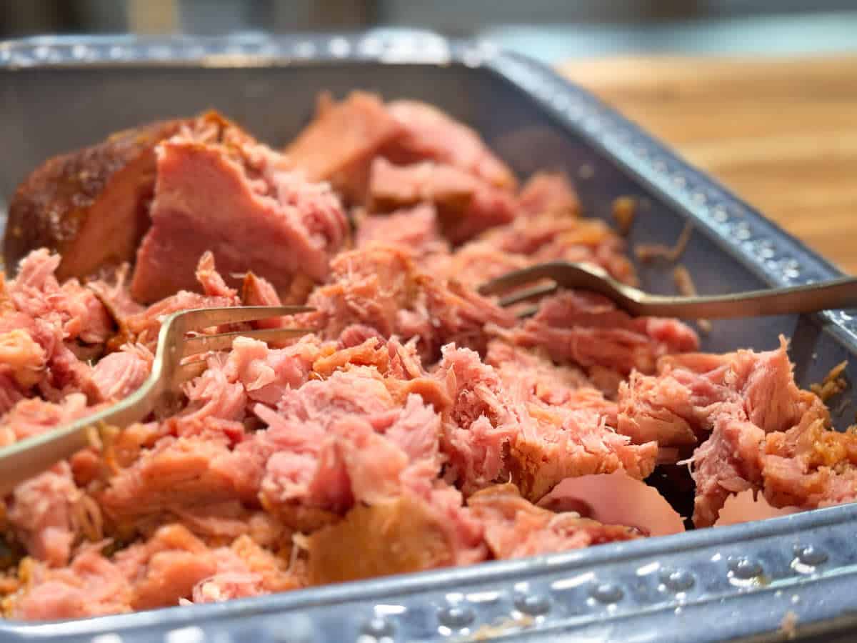A close-up of shredded pink meat, likely pulled pork, in a metal tray with two forks used for pulling apart the meat. The tray rests on a wooden surface in the background.