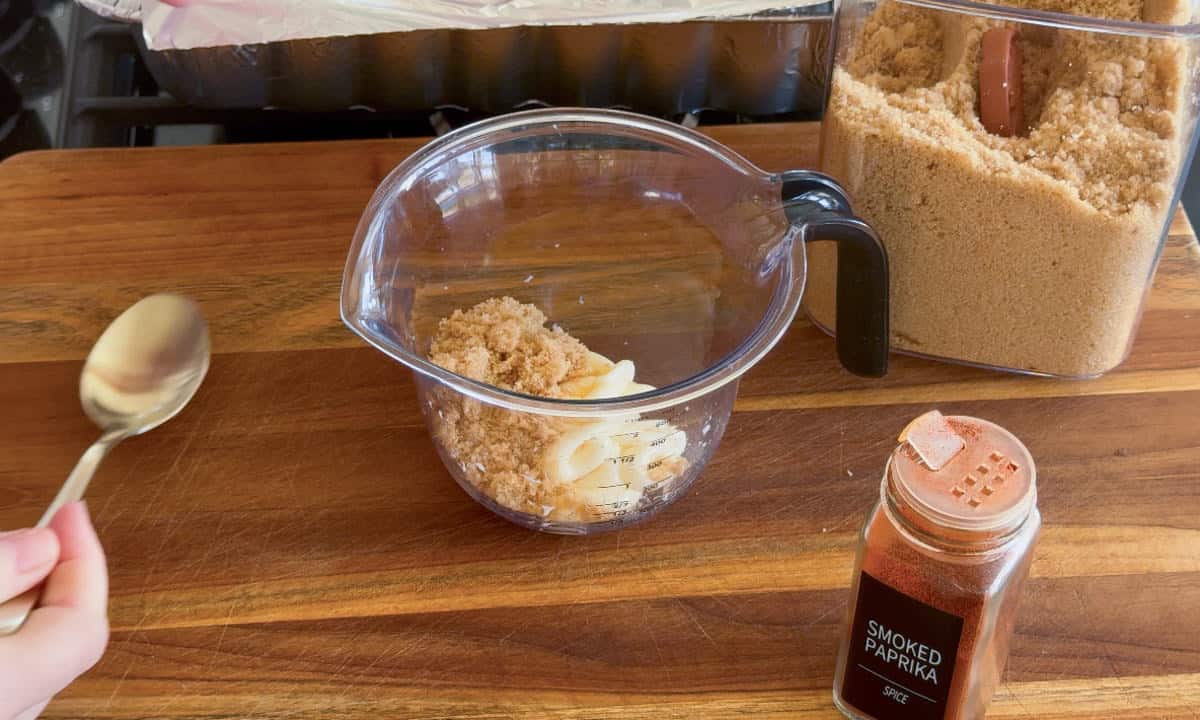 A hand holds a spoon near a measuring cup containing brown sugar and sliced butter on a wooden countertop. An open container of brown sugar and a jar of smoked paprika are also on the counter.