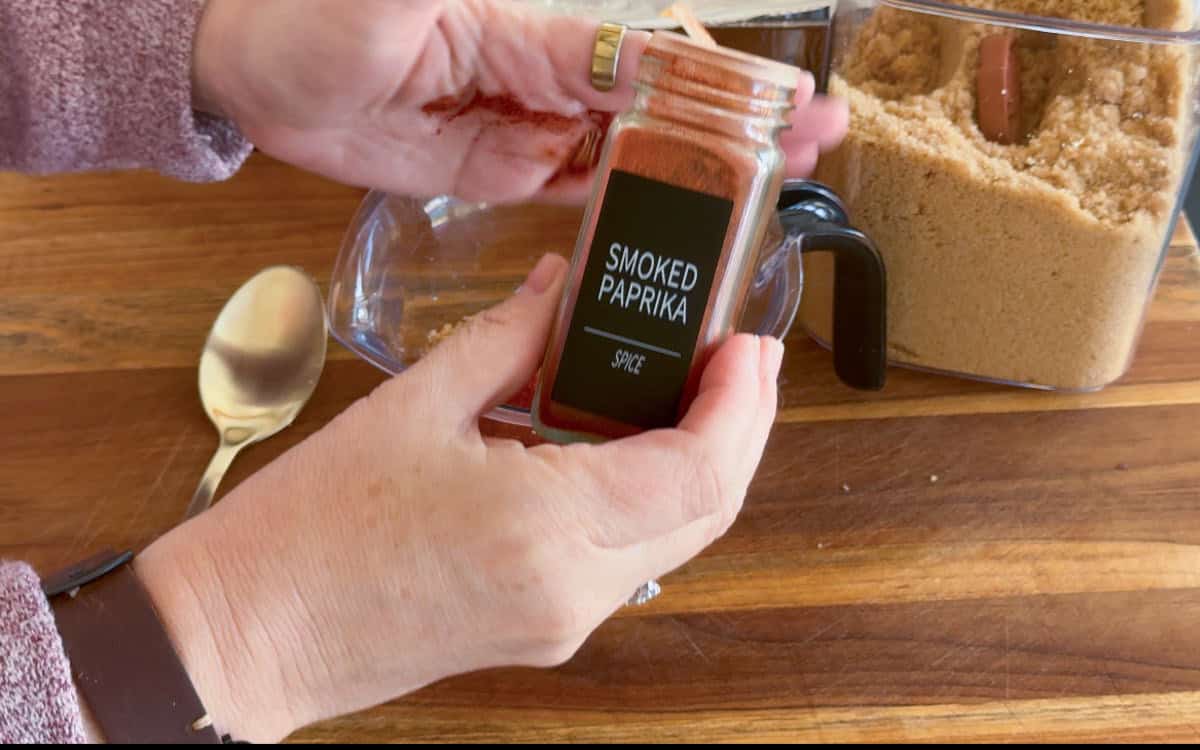 A person is holding a glass jar labeled "Smoked Paprika" above a clear bowl. There is a container of brown sugar, a wooden surface, and a metal spoon visible in the background.