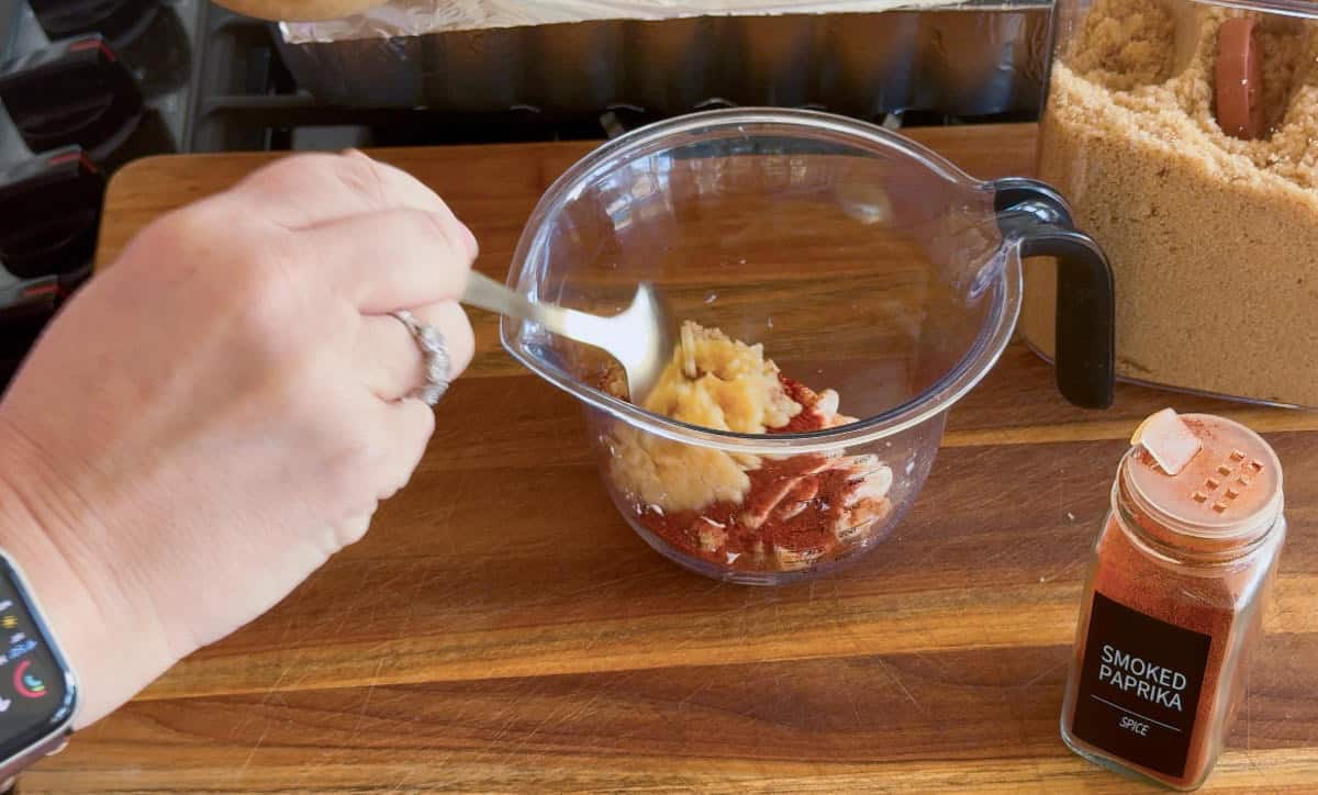A person mixes ingredients including smoked paprika and what appears to be minced garlic in a clear measuring cup on a wooden cutting board. A container of smoked paprika spice is visible next to the cup.