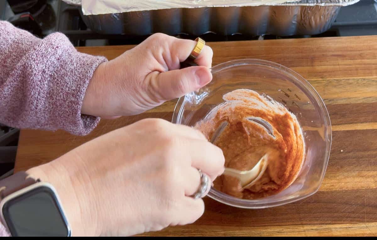 A person wearing a smartwatch and ring mixes a brownish paste in a small glass bowl with a spoon on a wooden cutting board. A foil-covered tray is visible in the background.