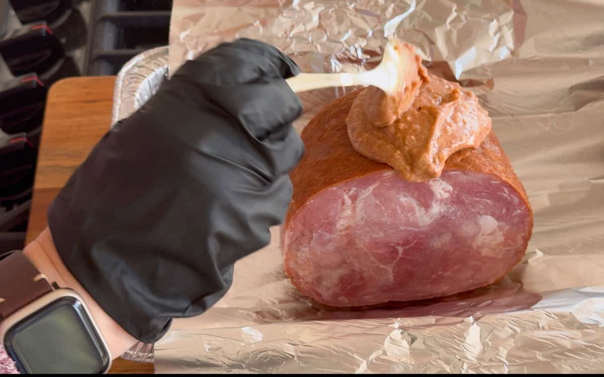 A person wearing a black glove applies a thick brownish paste onto a boneless ham using a wooden spatula. The ham rests on a sheet of aluminum foil. The person's wristwatch is visible.