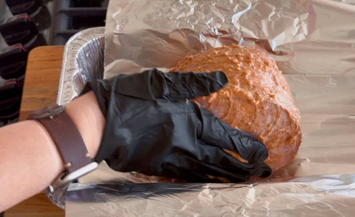 A person wearing a black glove and a watch is placing a seasoned meat roast onto a sheet of aluminum foil inside a foil tray.