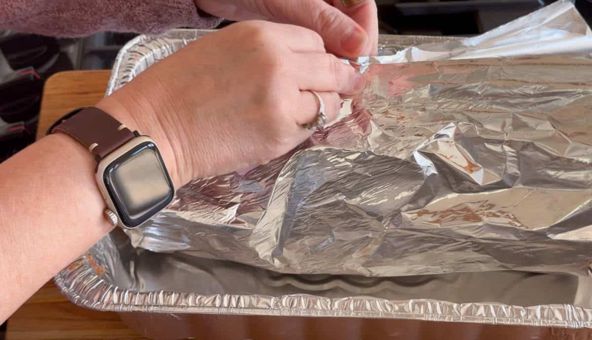 A person wearing a smartwatch and a ring covers a large rectangular aluminum pan with aluminum foil. The pan is on a wooden surface.
