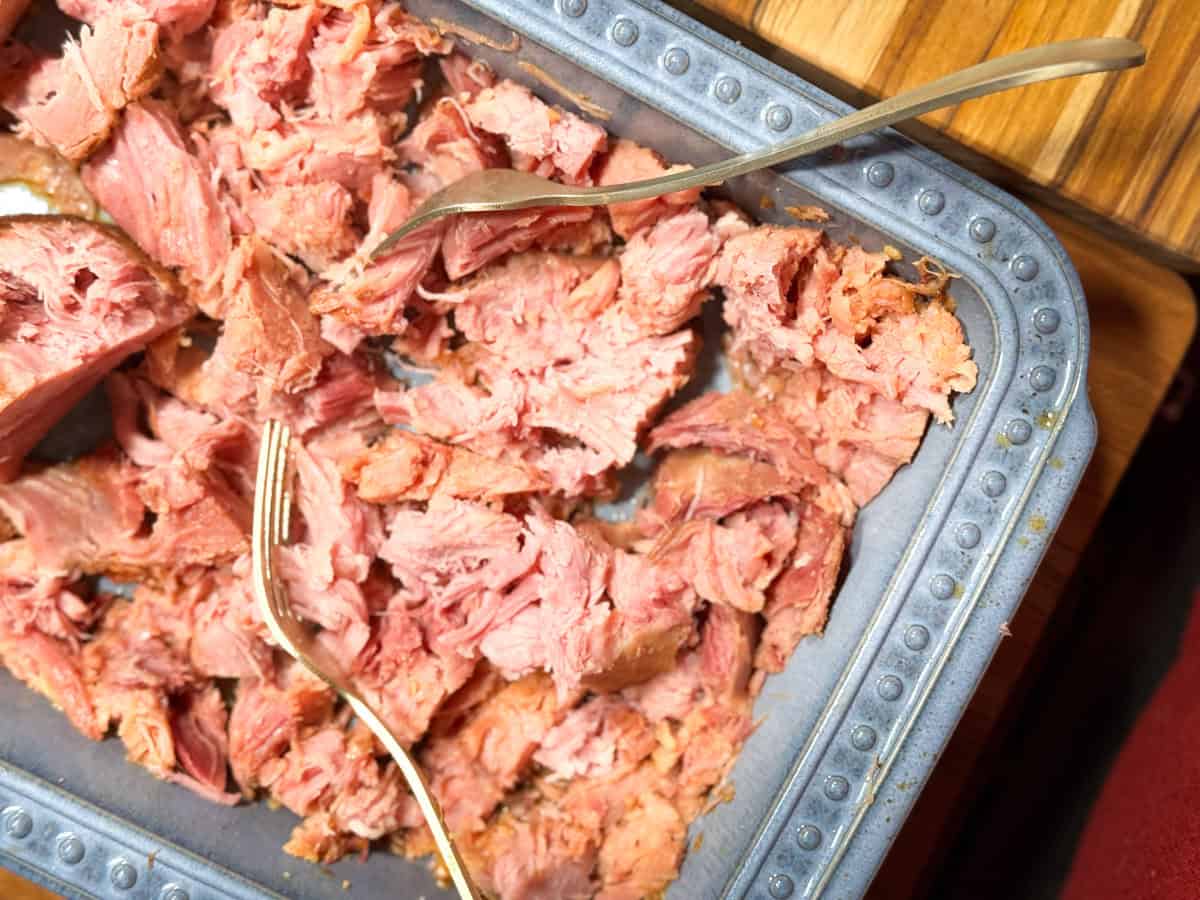A metal baking dish filled with shredded, cooked pink meat. Two forks are placed in the dish, positioned for pulling apart the meat. The dish rests on a wooden surface.