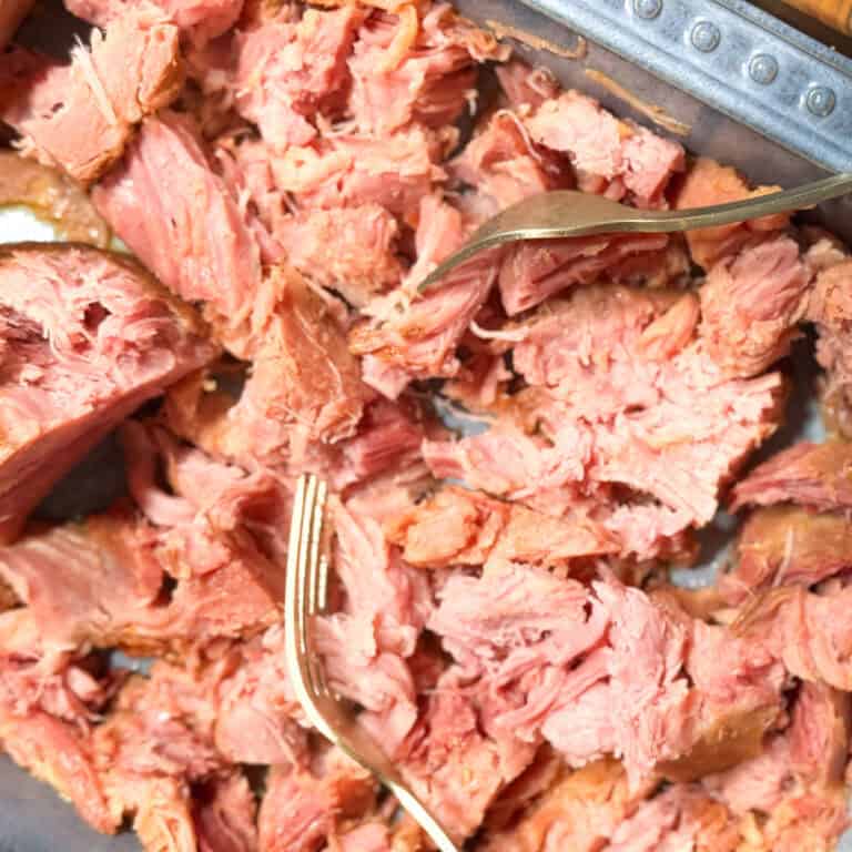 Shredded pink meat is being pulled apart with two forks in a metal pan. The meat appears tender and moist, with visible texture and fibers.