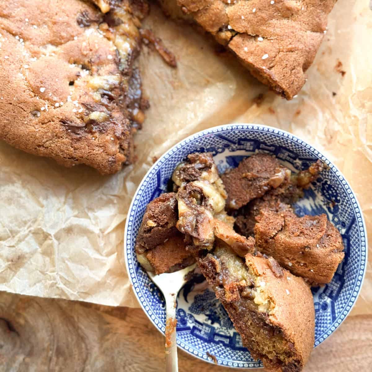 A blue and white bowl filled with chunks of a gooey chocolate chip cookie, placed on parchment paper with more cookie pieces nearby. A spoon rests in the bowl.