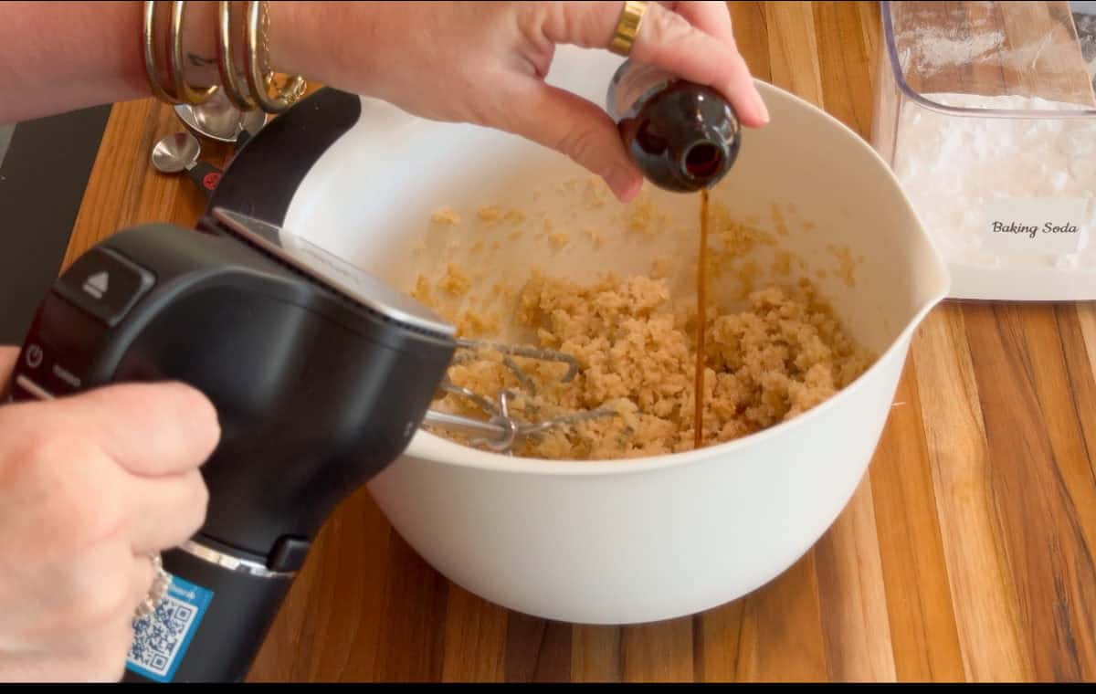A person pours vanilla extract into a white mixing bowl containing cookie dough while holding an electric hand mixer over the bowl. Measuring spoons and a baking soda container are on the wooden countertop nearby.