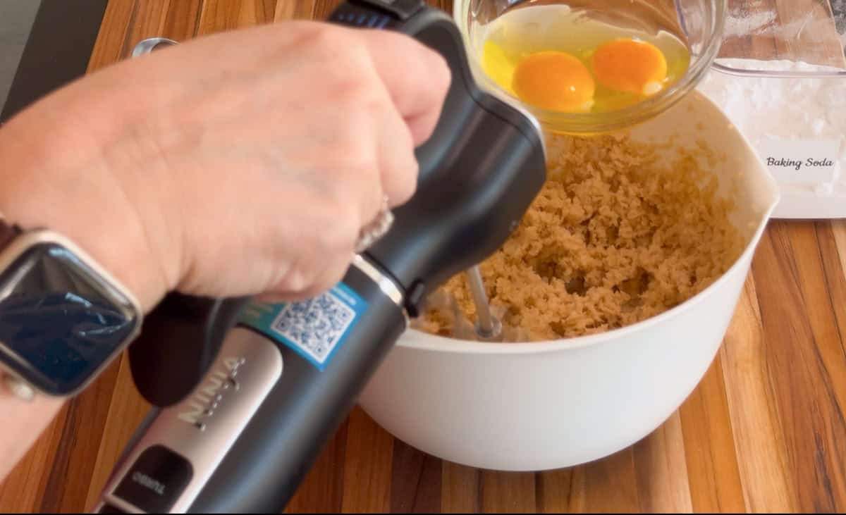 A person holding a hand mixer is about to add two eggs from a glass bowl into a white mixing bowl containing cookie dough. A container labeled "Baking Soda" is in the background on a wooden surface.