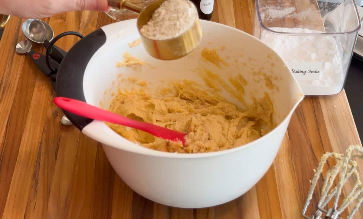 A person adds flour from a measuring cup into a mixing bowl filled with cookie dough, on a wooden countertop with a red spatula, baking soda container, measuring spoons, and mixer beaters nearby.